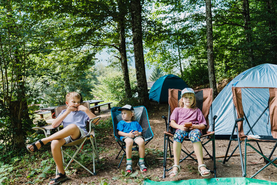 Three kids relaxing in camping chairs next to tents in a sunny green forest.