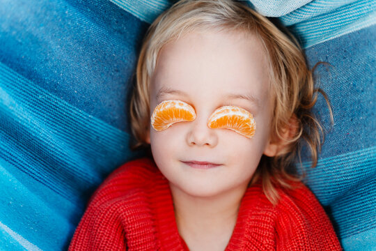 Funny child resting in a hammock holding mandarin slices over their eyes like orange glasses.