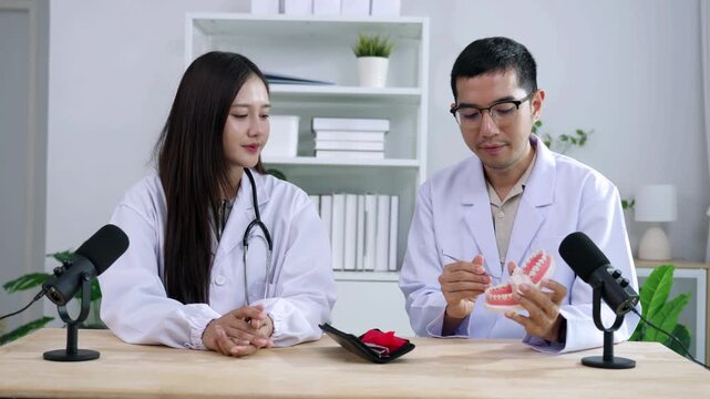 Asian male dentist holding a teeth model explaining oral care to a female doctor during a medical podcast interview. Recording a dental healthcare audio broadcast in a modern clinic studio.