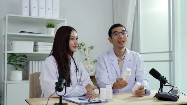 Asian male doctor explaining medicine from a blank white bottle to a female doctor during a medical podcast interview. Recording a healthcare audio broadcast in a clinic. Pharmacy concept.