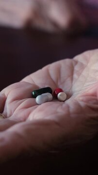 Senior person's wrinkled hand holding a variety of medicine pills and capsules, picking them up one by one for daily treatment, symbolizing healthcare and aging process in a close-up view