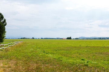 Naklejka premium Wide view of a lush green meadow with a wooden fence, distant mountains, and a cloudy sky.