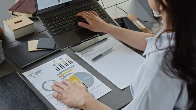 Gen Z girl analyzing financial charts and typing on laptop at office desk, tilt down shot. Footage is ideal for corporate training, business analysis, and financial planning marketing campaigns