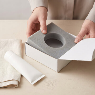 Hands pressing foam insert into cosmetic packaging carton beside matte white tube