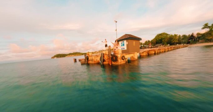FPV aerial view of a tropical coastline with calm turquoise water, a wooden pier extending into the sea, and lush palm trees under a soft sunset sky in Fiji.