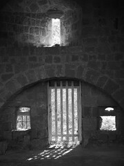 Fototapeta premium Black-and-white photograph of the interior of a historic, vaulted stone room with a barred entrance door and small, window-like openings through which daylight streams in, casting striking shadows on 