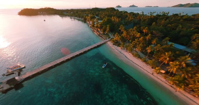 FPV aerial view of tropical island with a long wooden pier extending into turquoise waters surrounded by lush palm trees at sunset in Fiji.