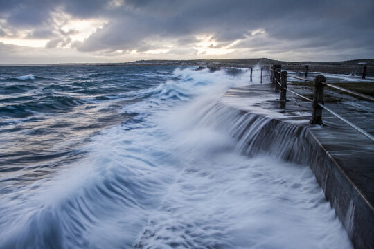 Ocean waves overflowing a concrete seawall, coastal landscape with dramatic weather and long exposure.