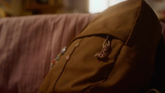 Brown school backpack with love keychain resting on pink sofa. Close-up shot is perfect for back-to-school marketing campaigns, teenage lifestyle content, or student accessory advertisements