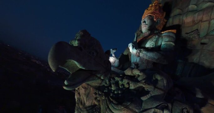 FPV aerial view of the massive Garuda Wisnu Kencana statue wing at dusk with city lights visible in the distance in Bali, Indonesia.