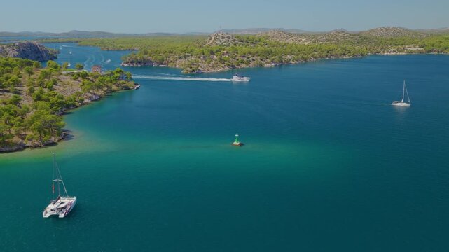 Aerial drone footage flying backward over Luka Channel near &Scaron;ibenik, Croatia, revealing turquoise waters, multiple boats and wakes, rocky shores, lush Mediterranean islands and scenic coastline.