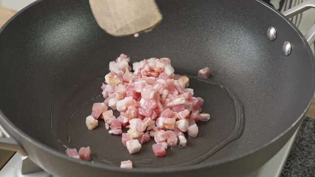 Cooking bacon/pancetta lardons in a non-stick wok. Real-time food preparation footage showing diced pancetta sizzling, rendering fat, and browning over medium heat in a kitchen pan.