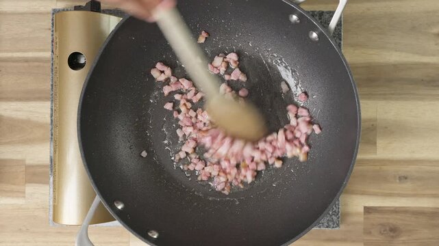 Cooking bacon/pancetta lardons in a non-stick wok. Real-time food preparation footage showing diced pancetta sizzling, rendering fat, and browning over medium heat in a kitchen pan.