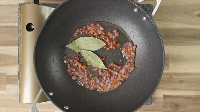 Adding bay leaves to bacon/pancetta lardons fryin in a non-stick wok. Real-time food preparation footage showing diced pancetta sizzling, rendering fat, and browning over medium heat in a kitchen pan.