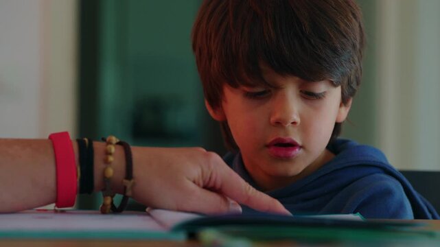 School-age boy reading aloud from an open workbook as an adult points to the page, concentrating on the lesson while seated at a table during home study indoors