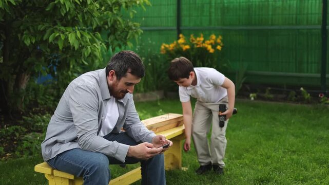 A man sits on a bench. His son is making something. A smartphone. Green grass. Slow motion video. High quality 4k footage