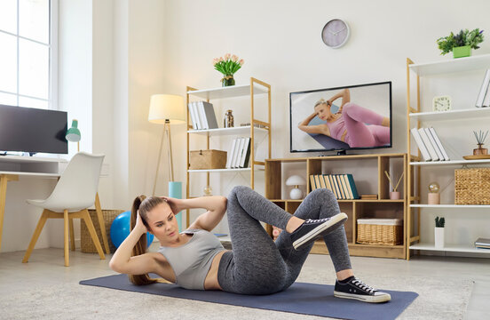 Woman home workout fitness exercise, online training. She does bicycle crunches on a mat in a living room, following a guided session on the TV. Clear concept of guided home fitness