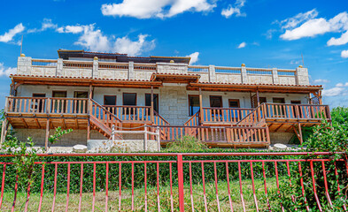 Fototapeta premium A two-story stone house features wooden balconies under a bright blue sky filled with white clouds.