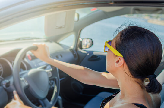 A young woman with sunglasses drives a car along a scenic coastal road during a sunny day.