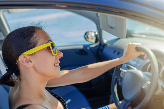 A cheerful woman wearing yellow sunglasses drives a car on a sunny day.