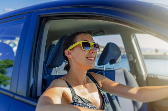 A smiling woman in yellow sunglasses drives a car on a sunny day.