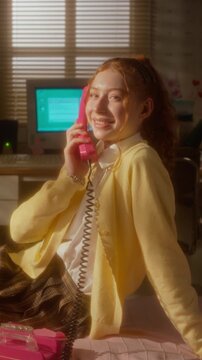 Vertical portrait of Caucasian teenage girl smiling while talking on pink retro corded telephone in 90s style bedroom. Footage perfect for nostalgia marketing campaigns