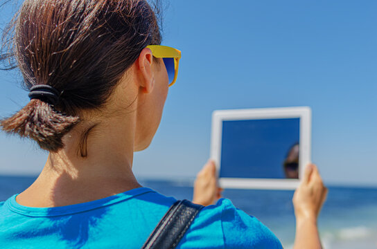 A woman wearing sunglasses holds a digital tablet outdoors near the blue sea under a clear sunny sky.