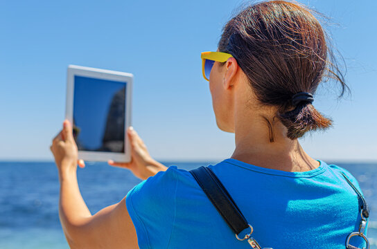 A woman in yellow sunglasses holds up a digital tablet to take a photo of the ocean on a bright sunny day.