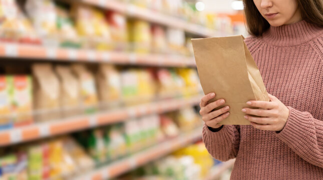 A woman stands in a grocery store holding a brown paper package. She examines the package closely while surrounded by colorful shelves filled with different products