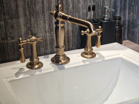 luxury interior photograph of a vintage antique brass widespread faucet set mounted on a white porcelain ceramic basin against a dark grey marble tile wall