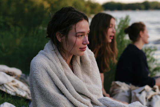 A group of young women shares a reflective moment by the riverside, wrapped in towels, showcasing the beauty of friendship and contemplative silence in nature's embrace.