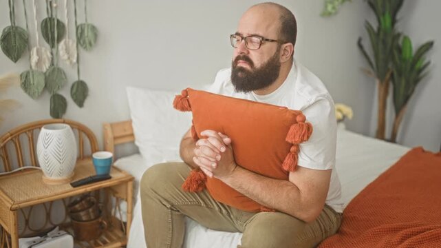 Bald bearded man clutches orange pillow on bed in building, hands interlaced and gaze turned aside, seated near bedside table and plants; anxiety.