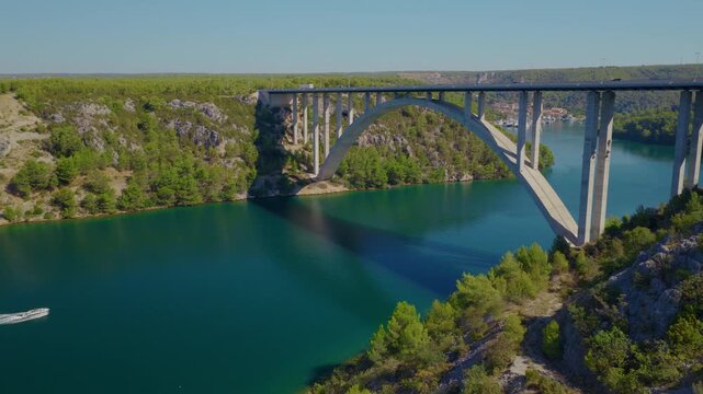 Aerial drone view of Krka Bridge in Croatia, with a smooth orbital camera movement around the concrete bridge above turquoise river waters, rocky landscape and lush Mediterranean vegetation.