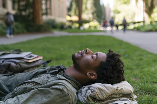 A young man lies peacefully on the grass, eyes closed and surrounded by greenery, embodying relaxation and a deep connection with nature during a sunny day.