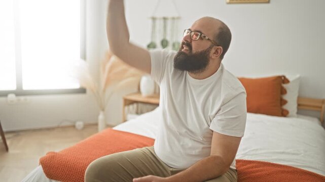 Man reaches hand upward while seated on bed in bedroom, head tilted and eyes fixed upward toward ceiling; quiet contemplation.