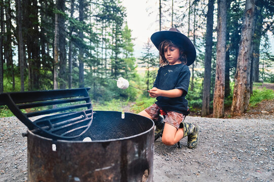 Boy in a cowboy hat crouching beside a campfire holding a marshmallow stick