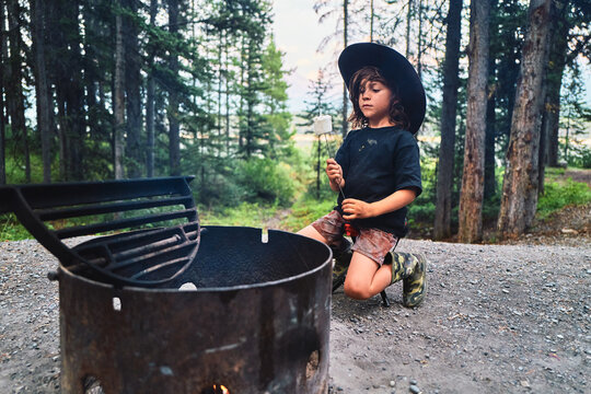 Boy in a cowboy hat looking at a roasted marshmallow beside a fire pit