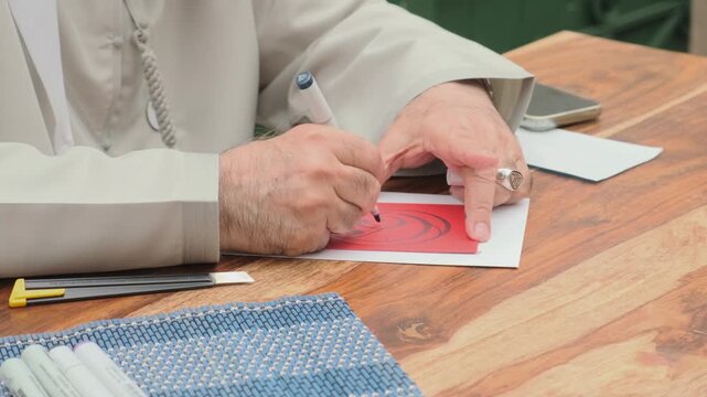 Arabic calligraphy hand writes ornate curves on red paper with careful pen pressure. Robed hands guide each stroke over wooden table beside pens and tools for focused script practice.