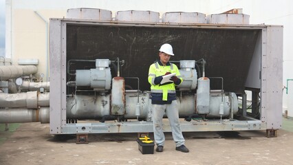 Fototapeta premium Asian male technician in hardhat checking rooftop industrial cooling tower and pipes with clipboard. Commercial HVAC refrigeration maintenance, technical system audit and industry concept.