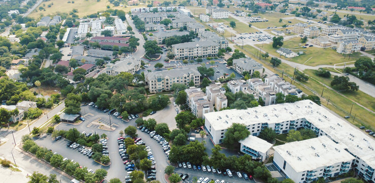 Modern apartment buildings border Mahncke Park residential district. Winding parking lanes host parked vehicles amidst lush trees, multi family units, open fields, San Antonio, Texas