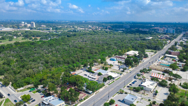 Dense woodlands border commercial storefronts along Broadway near Brackenridge Park. Paved parking spaces, low rise structures, open roadways dominate lively urban landscape, San Antonio, Texas
