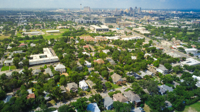 Distant city skyscrapers rise beyond Mahncke Park residential district. Dense tree foliage surrounds single family bungalows, apartment courts, paved access drives, San Antonio, Texas