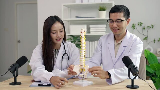 Asian female doctor pointing at a spine model explaining orthopedics to a male doctor during a medical podcast interview. Recording a healthcare audio broadcast in a modern clinic studio.