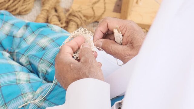 Close up hands repair netting with steady motion and precise knots. Elderly fisherman in white and blue clothing practices arabic craft linked to heritage and daily labor through careful handwork.