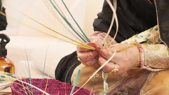 Skilled hands weave palm frond strips into basket form with careful rhythm. Gold jewelry and embroidered dress suggest Emirati heritage, while colored fibers fan outward in traditional Arab craft.