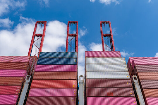 Low angle view of colorful stacked cargo shipping containers with large red gantry cranes against blue sky, global logistics and maritime trade concept