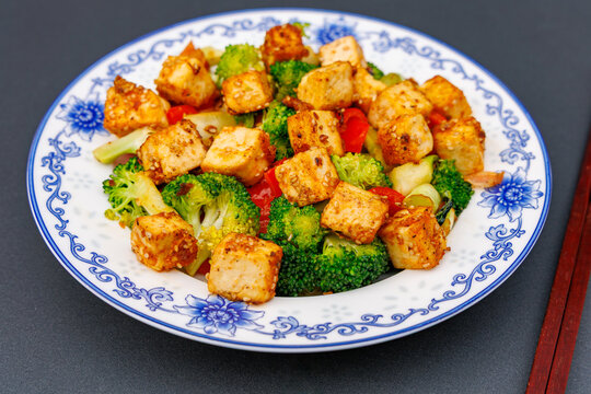 Crispy fried tofu stir fry with broccoli and red bell pepper on blue and white porcelain plate with chopsticks, asian vegan cuisine concept