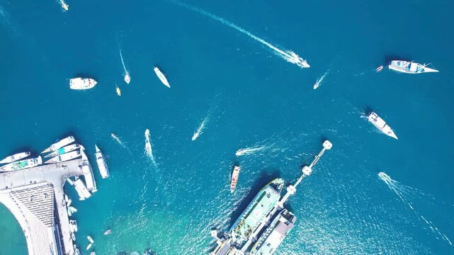 Top down drone footage of Labuan Bajo harbor in Flores, Indonesia, showing passenger ferries at the pier, speedboats and small boats moving across tropical blue water with visible boat