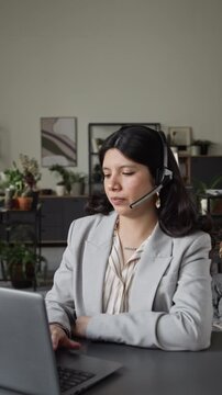 Vertical video of Hispanic young adult woman wearing headset, talking and typing on laptop. Clip is ideal for customer support, remote work, and online consulting advertisements