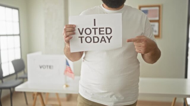 Man holding i voted today paper sign, pointing finger to sign inside building polling station next to small american flag; civic pride.